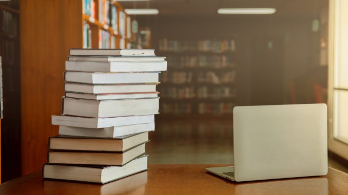 Stack of old books and laptop computer on desk in library background.