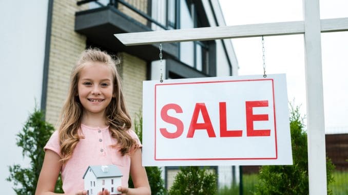 Cheerful kid holding carton house model near board with sale letters
